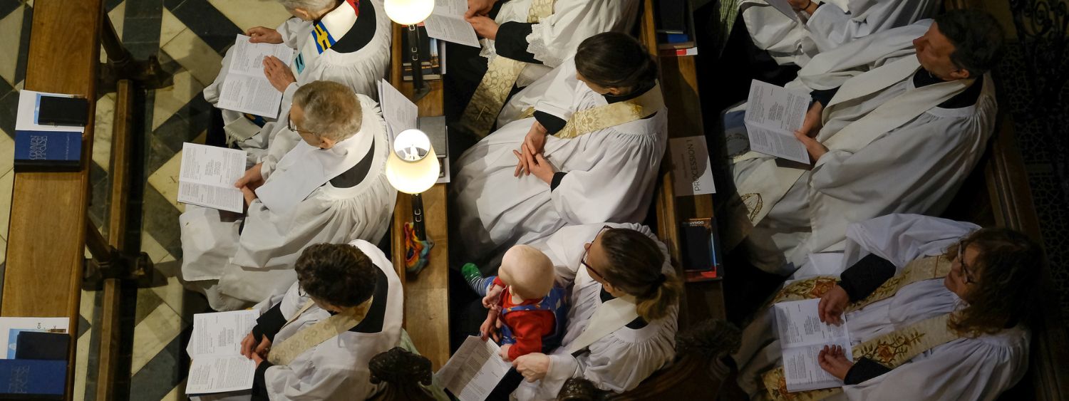 Aerial view of clergy sitting in pews during a service. One woman holds a baby on her lap.