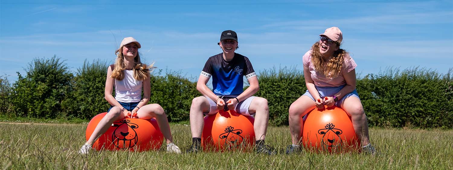 Three teenagers bounce on space hoppers in a sunny field, laughing at the camera