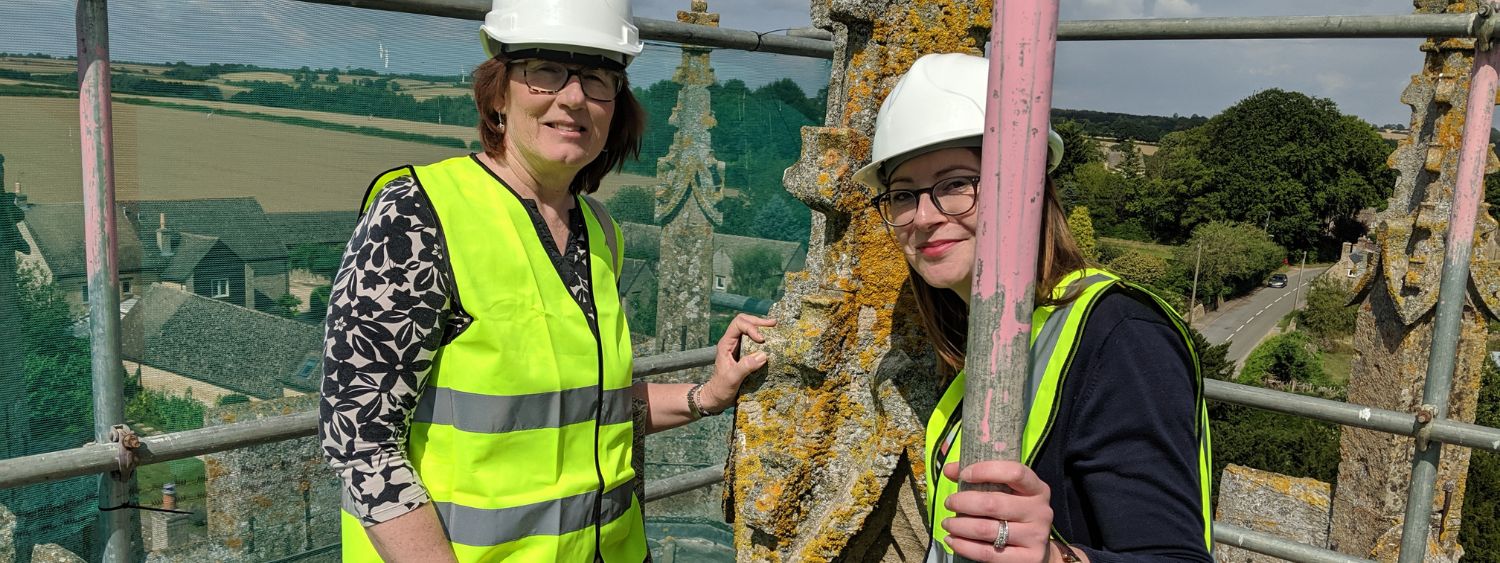 Two women in hi vis jackets and hard hats stand on scaffolding at the top of a church roof