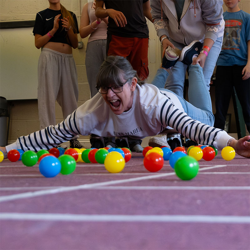 A woman lies face down on a skateboard, arms outstretched as she is wheeled towards a pile of colourful plastic balls. Her face is a picture of glee
