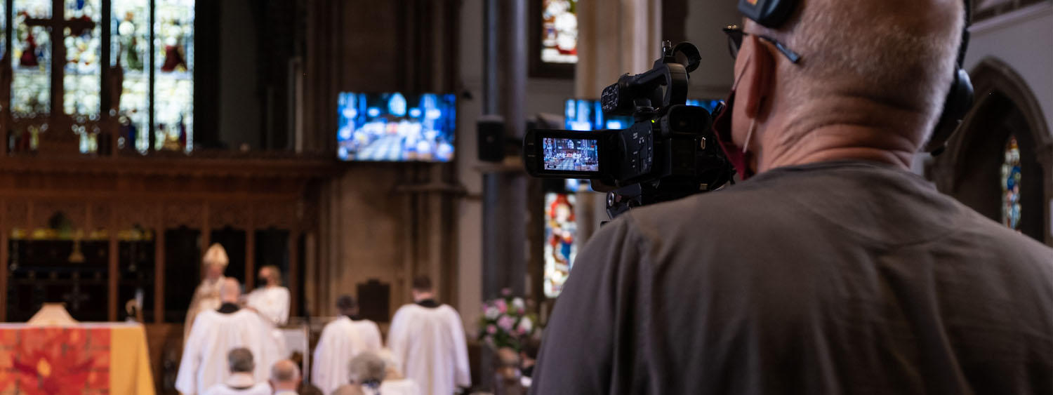 Over the shoulder photo of a man wearing headphones, aiming a large video camera at a church service