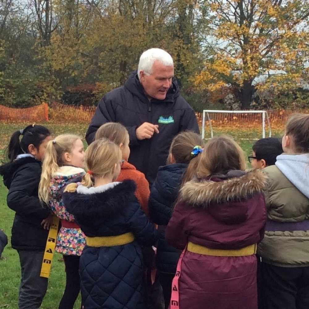 Chris Andrew is standing on a football field with a goal behind him talking to a group of primary school children wearing coats and tag rugby waist bands.