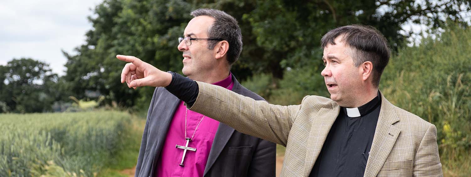The Revd Neil stands next to Bishop Gavin on a country road. He is pointing into the distance, and Bishop Gavin is following his gaze.