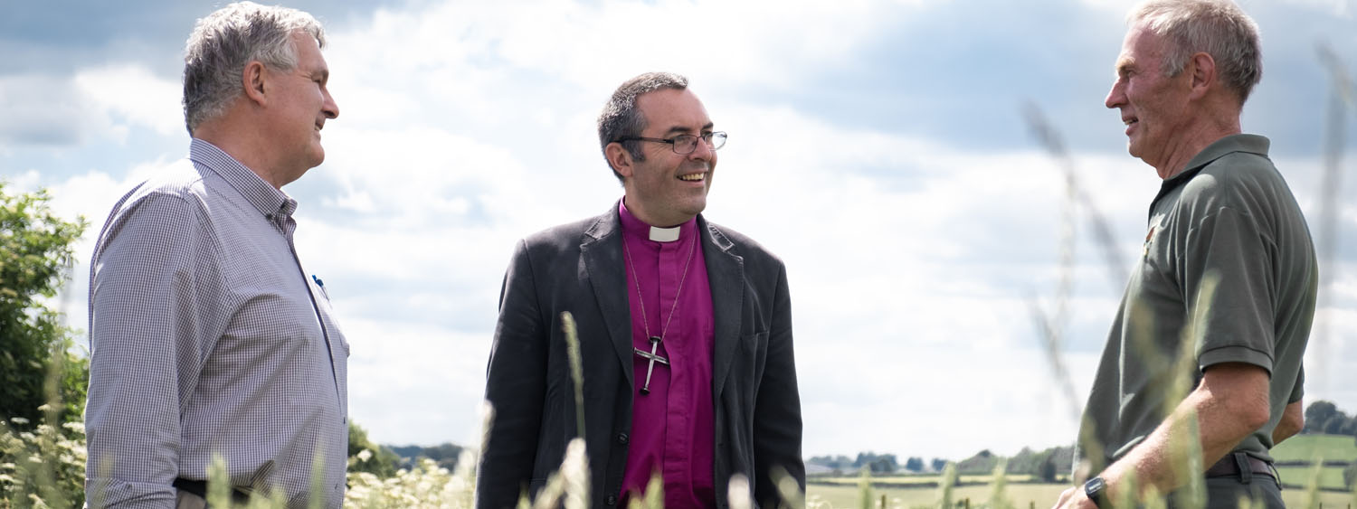 Bishop Gavin talks happily to two farmers in a field of tall grass