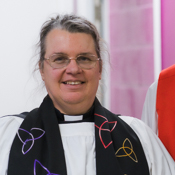 The Revd Hannah Lewis is robed wearing a clerical collar and black stole with coloured patterns. Hannah is wearing glasses and smiling.