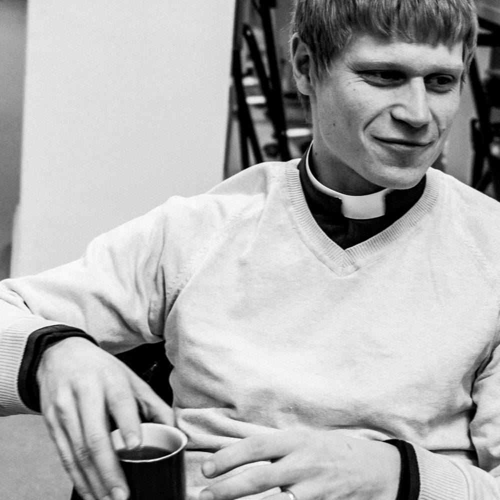 Black and white photo of the Revd Rob Glenny, smiling and hold a cup of coffee. He is wearing a shirt and clerical collar and a light coloured v neck jumper.