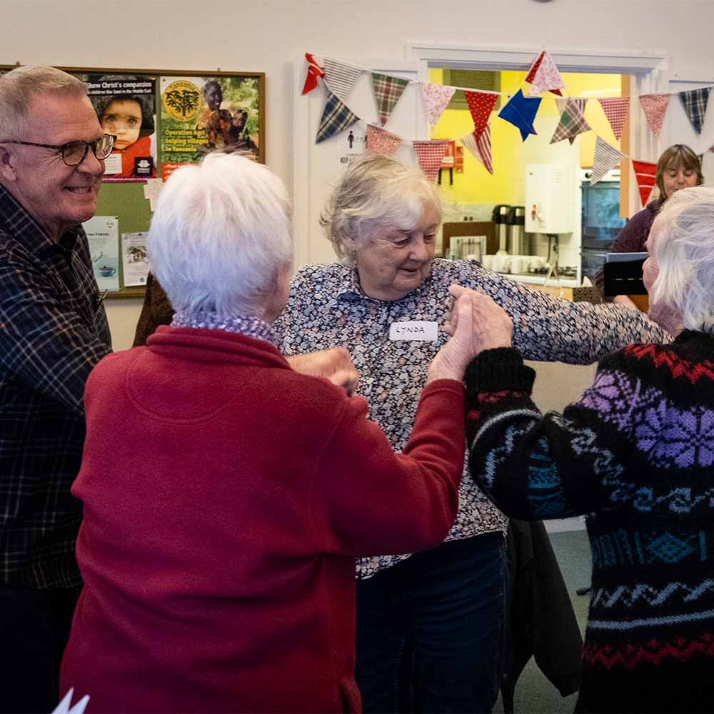 A group of four elderly people hold hands and dance together in a circle. The room they are in is decorated with colourful bunting