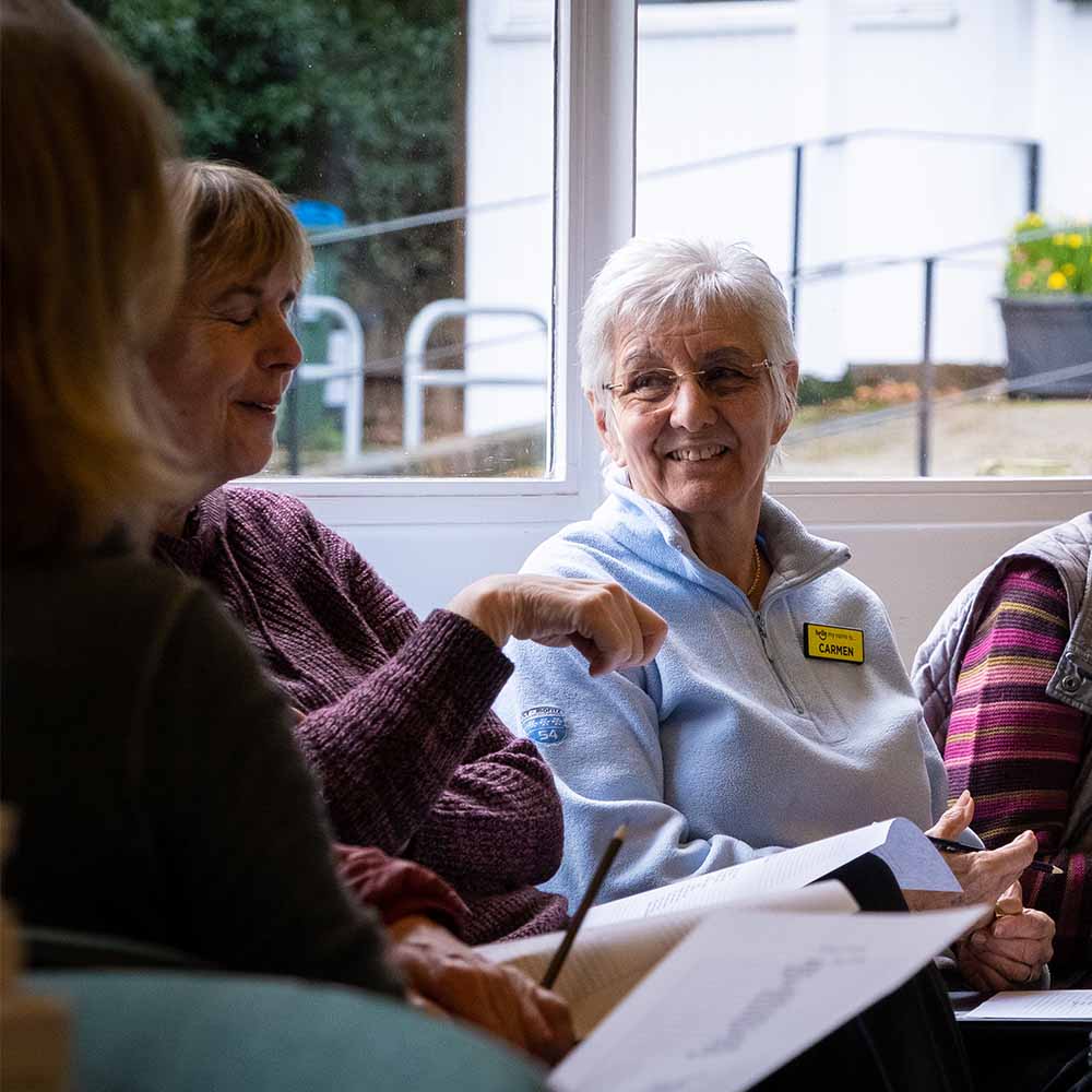 A group of people sit in a circle talking. A woman is looking to her right and smiling at the woman speaking