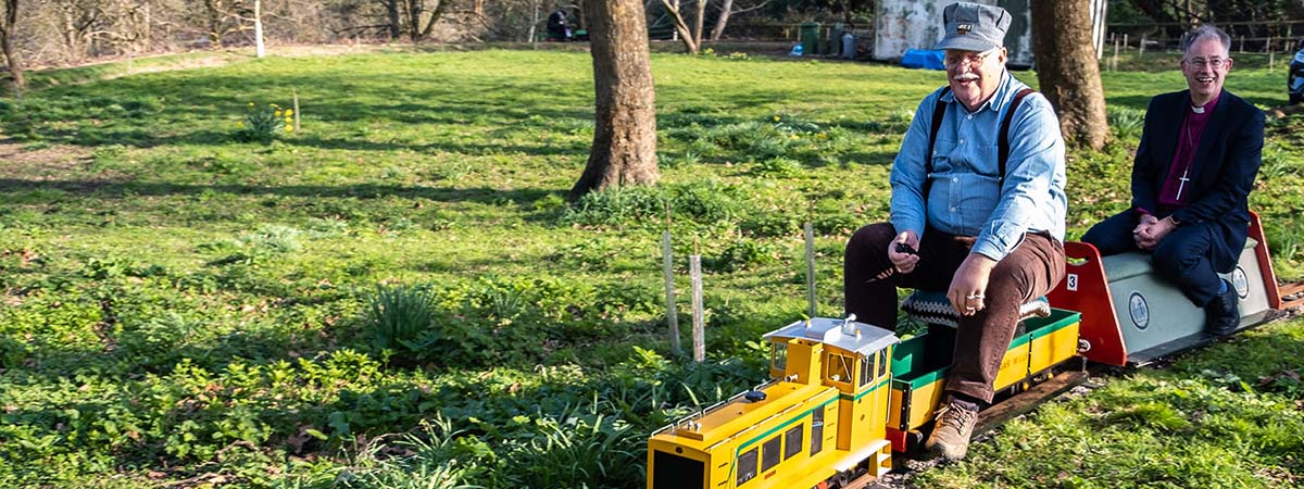 Bishop Steven smiles as he rides on the last carriage of a tiny electric train. The train driver sits in front. The train is yellow and the track goes through greenery and trees