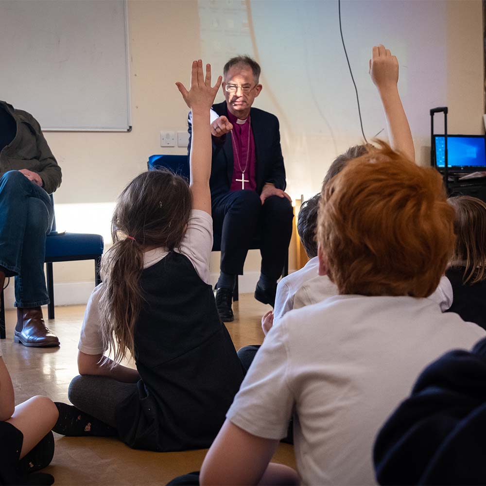 Bishop Steven sits in a church hall looking towards school children sat on the floor. The children have their hands stretched into the air and Bishop Steven is pointing forward at one girl with her hand up