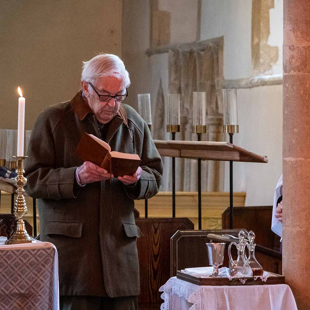 A man stands at an altar reading the bible. On the altar in front of him is a lit taper candle in a gold candlestick