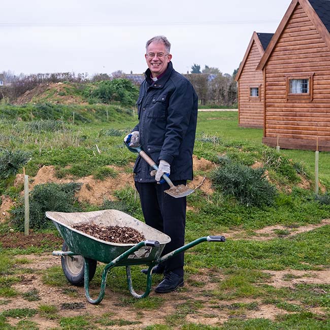 Bishop Steven stands in a field holding a spade over a wheelbarrow full of bark. He is looking at the camera and smiling. In the background are some wooden huts.