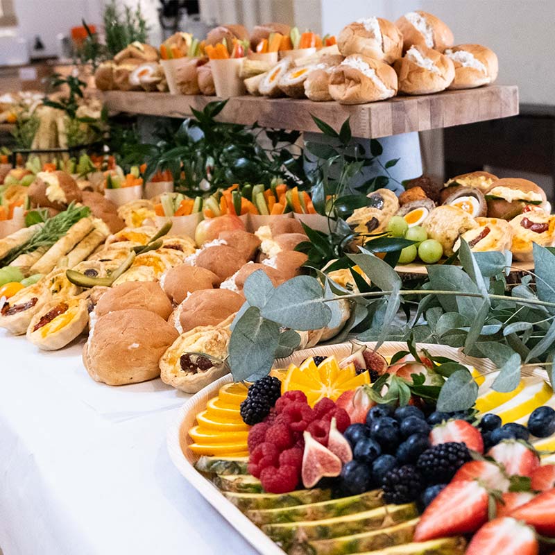 A decadent buffet spread on a long table, with rolls, fruit platters, tarts and greenery