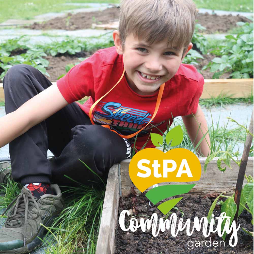 A young boy sits outside on the floor in front of a flowerbed, grinning up at the camera. The St Peter's Aylesbury Community Garden logo is overlayed on the bottom right 