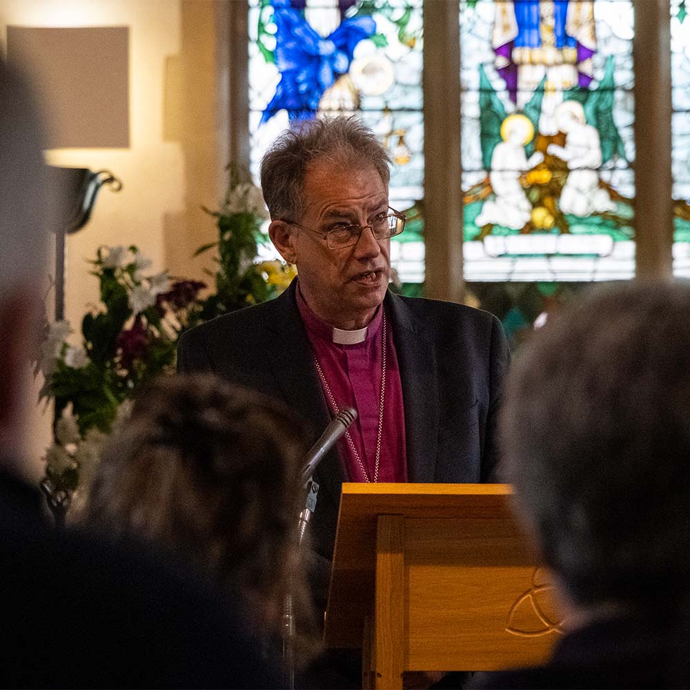 Bishop Steven preaches from a lecturn. Behind him is a large stained glass window. Out of focus in the foreground are members of clergy making up the congregation