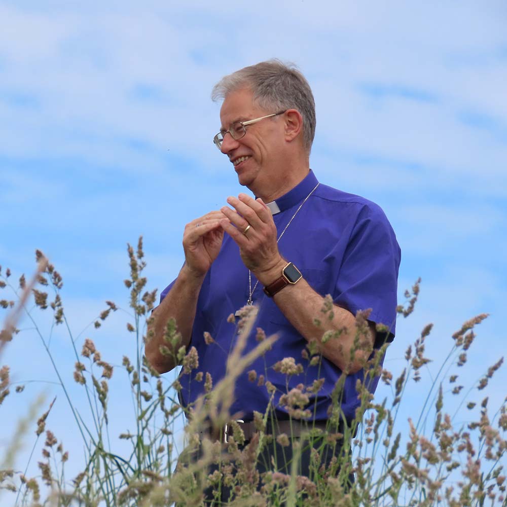 Bishop Steven stands in a field of tall grass on a clear sunny day. He is smiling and clapping at something out of view