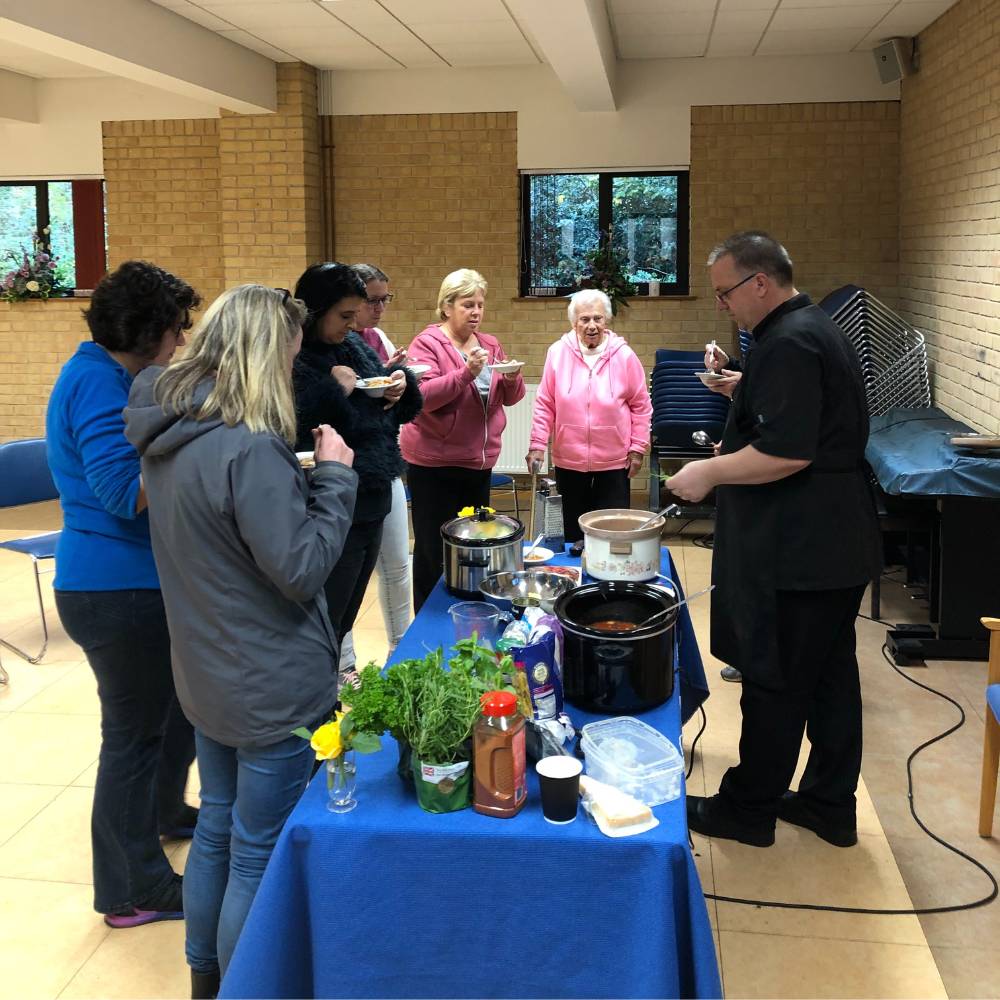 group of people standing round a table with food and a slow cooker, a chef is showing them how to use the cooker and different recipes.