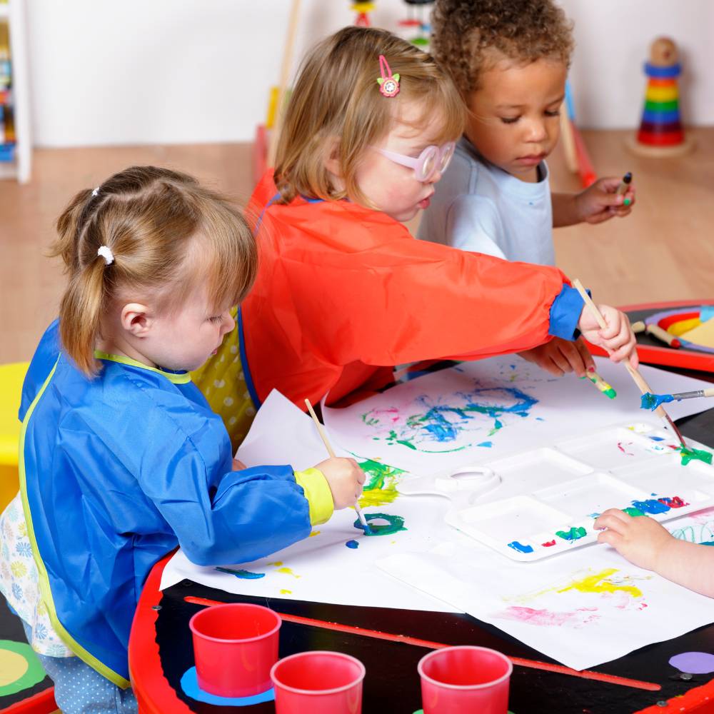 Three toddlers wearing aprons and sitting at a small table painting with brushes and different coloured paint.