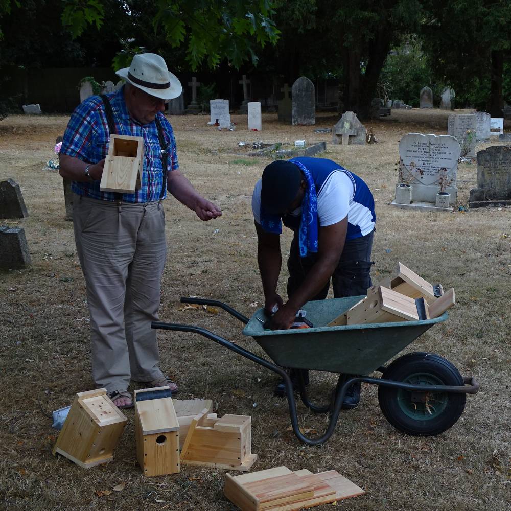 Two men with multiple newly built wooden bird boxes in a wheelbarrow in St Peter's churchyard.