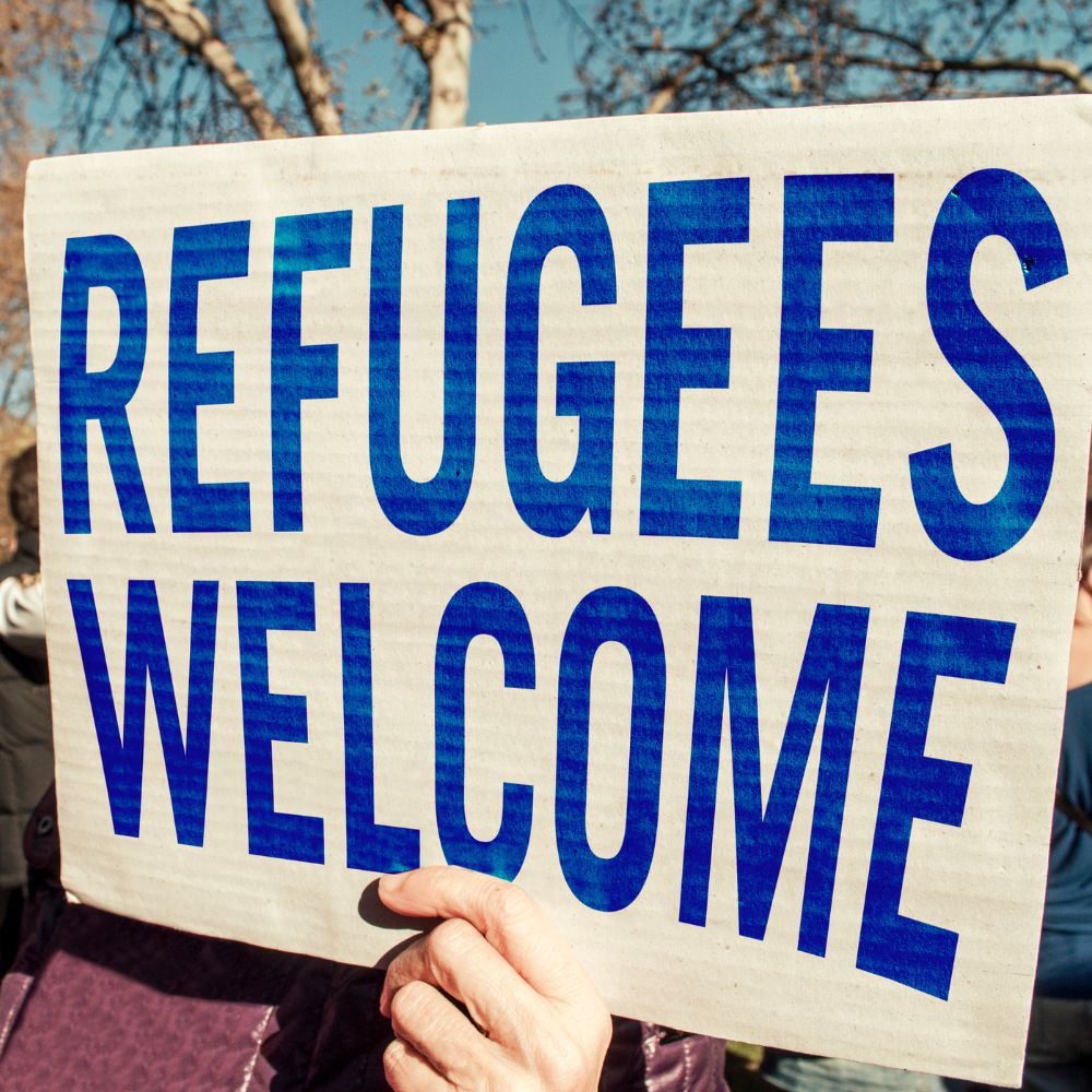 Handmade sign reading "REFUGEES WELCOME" written in blue.