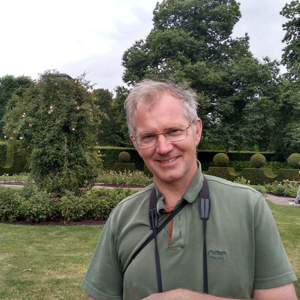 David is stood in a large garden, he is wearing a green polo shirt and is smiling.