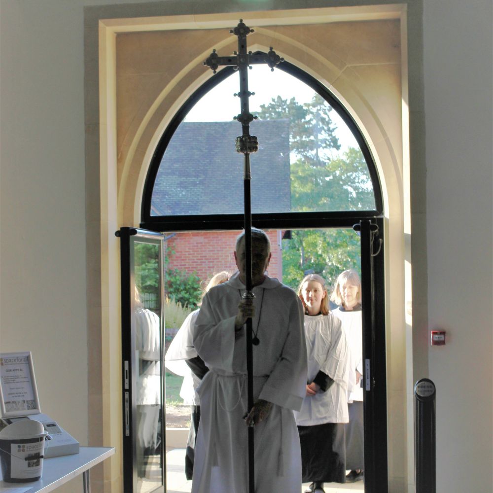 New glass ground level double entrance door at All Saints Church, a verger carries a crucifix into the church with a robed procession of people behind.