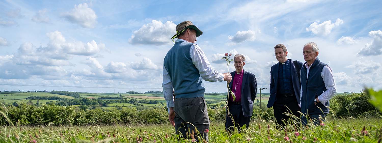 Bishop Steven, a vicar and another man stand looking towards a farmer holding out a large flower in a field. In the background are acres of farmland
